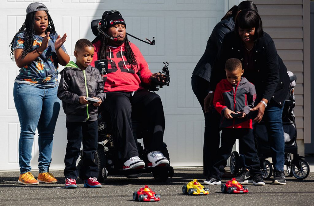 a family of five playing with r/c cars. Two of the cars are being controlled via remote control by two able bodied boys. The third is being controlled by a man in a wheelchair with a headset that has a pipe. Sipping and puffing into the pipe controls the r/c car.