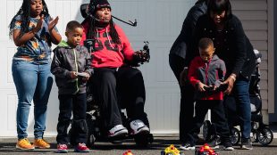 a family of five playing with r/c cars. Two of the cars are being controlled via remote control by two able bodied boys. The third is being controlled by a man in a wheelchair with a headset that has a pipe. Sipping and puffing into the pipe controls the r/c car.