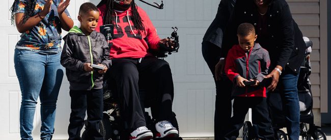 a family of five playing with r/c cars. Two of the cars are being controlled via remote control by two able bodied boys. The third is being controlled by a man in a wheelchair with a headset that has a pipe. Sipping and puffing into the pipe controls the r/c car.