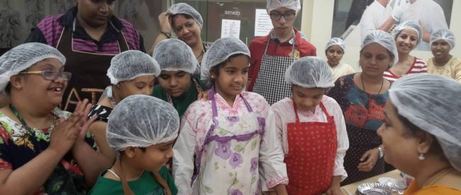 a group of children inside a kitchen listening to baking instructions from an adult mentor.