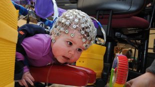 An infant shown wearing an electrodes studded cap. A second person is holding a toy in front of her, to encourage her to move towards the toy.