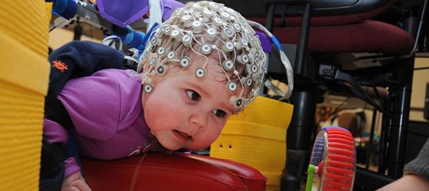 An infant shown wearing an electrodes studded cap. A second person is holding a toy in front of her, to encourage her to move towards the toy.