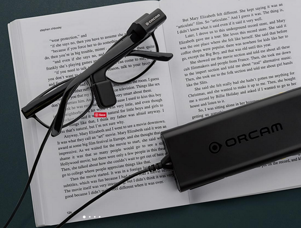 a pair of glasses with orcam myeye attached to the right stem resting on a book