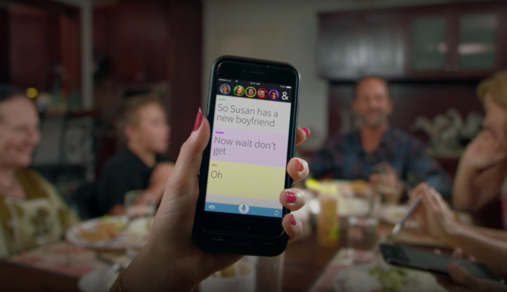 A person shown holding their phone at the dinner table. The phone shows Ava showing all the dialogues of people at the dinner table.