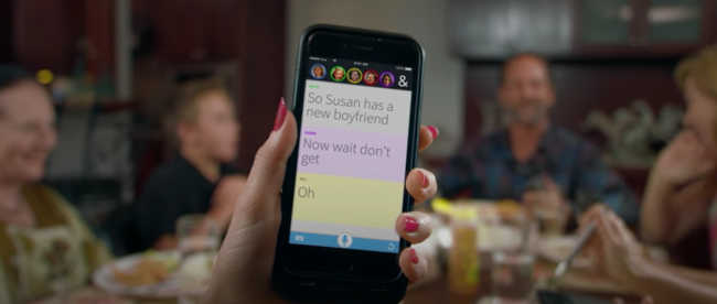 A person shown holding their phone at the dinner table. The phone shows Ava showing all the dialogues of people at the dinner table.