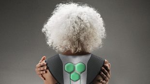 Photo of the back of an elderly person with wavy white hair. She is seen wearing a soft exoskeleton and holding both her arms.