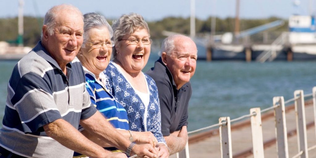 a group of four seniors standing near a lake, looking at something and laughing.