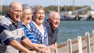 a group of four seniors standing near a lake, looking at something and laughing.