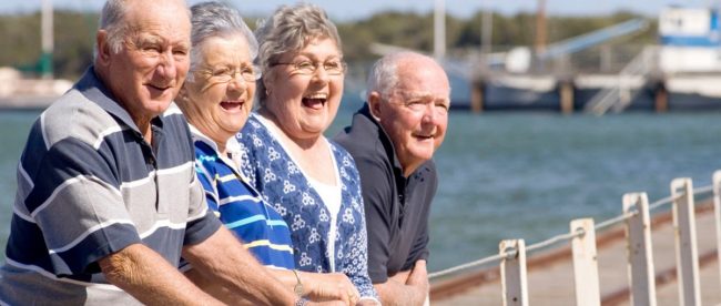 a group of four seniors standing near a lake, looking at something and laughing.