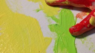 a child's hand seen covered in red paint and resting on a canvas that has yellow and green paint smudged on it.