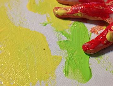 a child's hand seen covered in red paint and resting on a canvas that has yellow and green paint smudged on it.