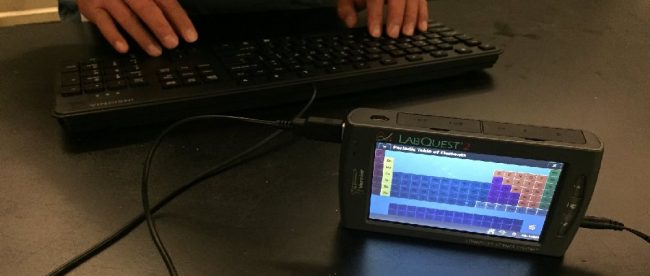 a keyboard is connected to a labquest 2, and the periodic table is displayed on the labquest 2's color screen. A person's hands are also placed on the keyboard.