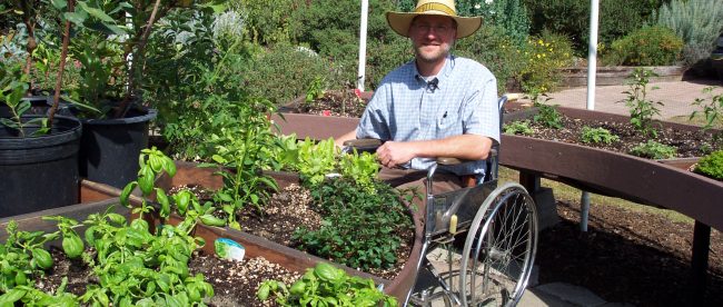 A man wearing a hat is seen in his wheelchair. In front of him are some plants. It looks like he is in a garden.