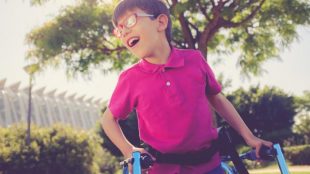 A child wearing a megenta shirt and glasses seen holding a walker. He is smiling.