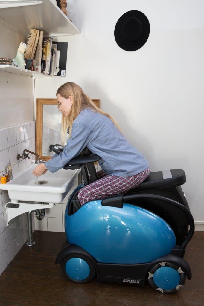 A woman seen sitting in the bathroom sitting in her Rodem mobility chair. She is leaning in to the wash basin and washing her hands.