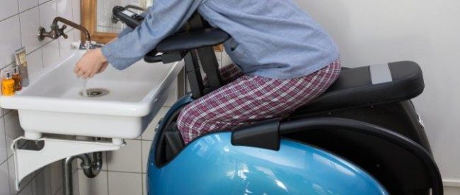 A woman seen sitting in the bathroom sitting in her Rodem mobility chair. She is leaning in to the wash basin and washing her hands.