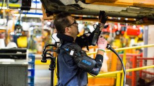 a person seen working on a car in an assembly line in a car manufacturing plant. This person is wearing the eksovest, and is seen drilling into the car body that is in an overhead position.