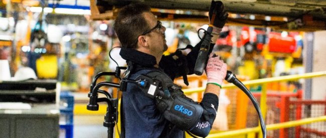 a person seen working on a car in an assembly line in a car manufacturing plant. This person is wearing the eksovest, and is seen drilling into the car body that is in an overhead position.