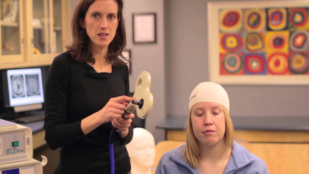 dr. gillick seen here with a patient. She is holding an equipment in her left hand. The patient is wearing a swim cap with markings of the spots in her brain that cause movement.