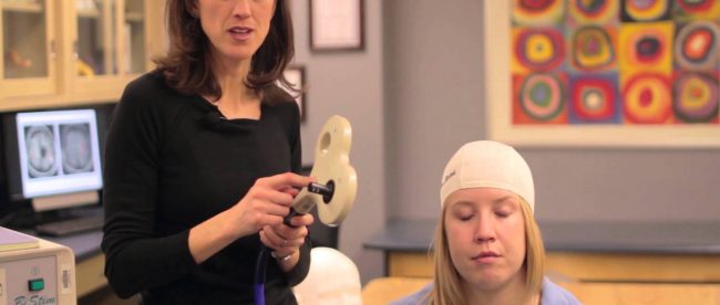 dr. gillick seen here with a patient. She is holding an equipment in her left hand. The patient is wearing a swim cap with markings of the spots in her brain that cause movement.