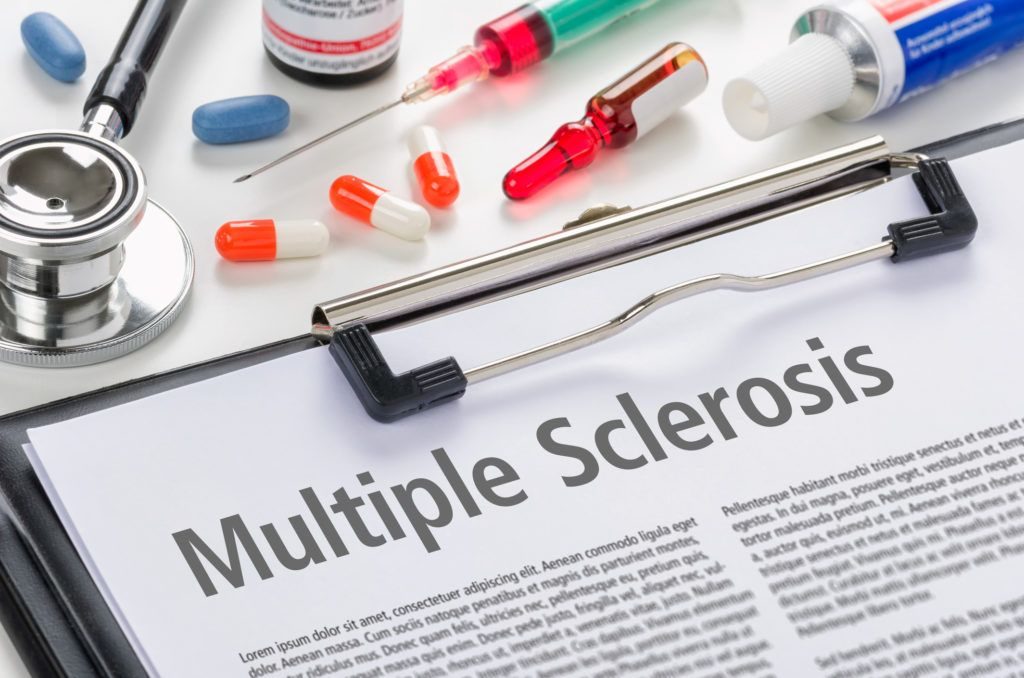 a clipboard with a sheet titled multiple sclerosis. A bunch of pills and a syringe are seen right next to it.