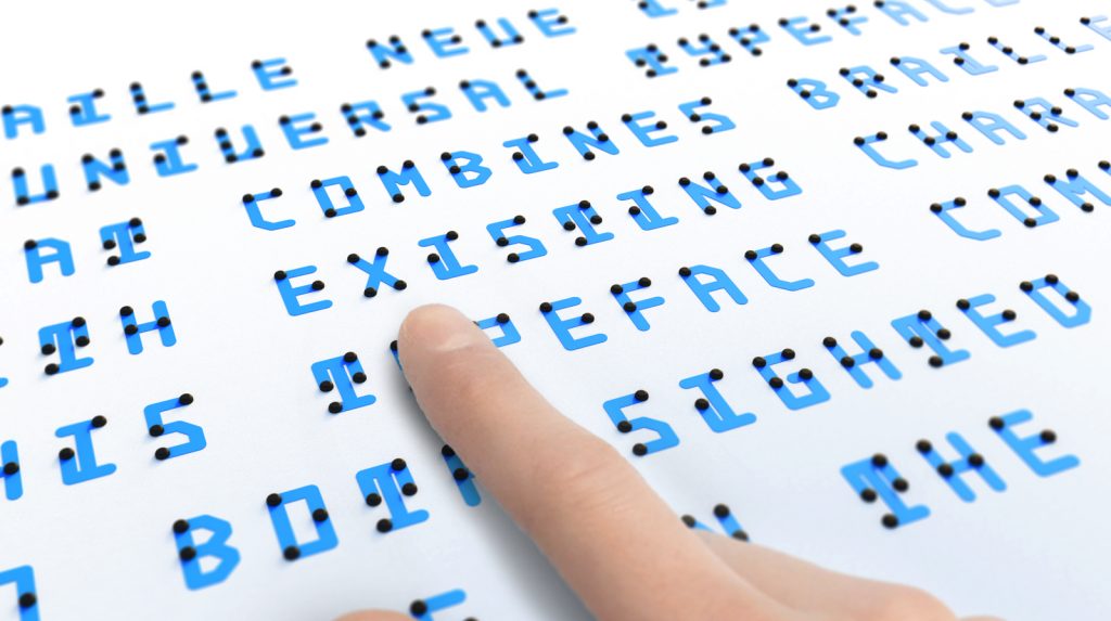 Close up of text in Braille Neue. A finger is seen on the text presumably of a blind person feeling the Braille bumps.