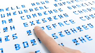 Close up of text in Braille Neue. A finger is seen on the text presumably of a blind person feeling the Braille bumps.