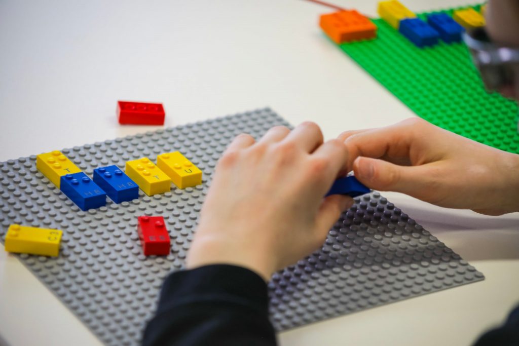 close up of a child's hands shown playing with braille lego blocks. Braille legos will have letters, numbers and punctuation printed on them as well so both sighted and blind children can play together.