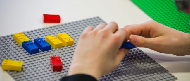 close up of a child's hands shown playing with braille lego blocks. Braille legos will have letters, numbers and punctuation printed on them as well so both sighted and blind children can play together.