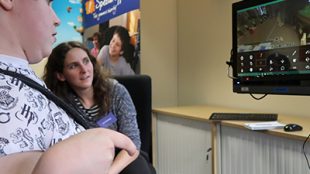 becky seen playing minecraft with her eyes. There is another person sitting next to her watching her gameplay on the monitor.