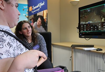 becky seen playing minecraft with her eyes. There is another person sitting next to her watching her gameplay on the monitor.