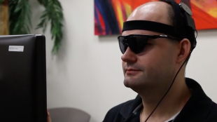 a person with acquired blindness is seen wearing sunglasses with camera attached to them. He is sitting in front of a computer.