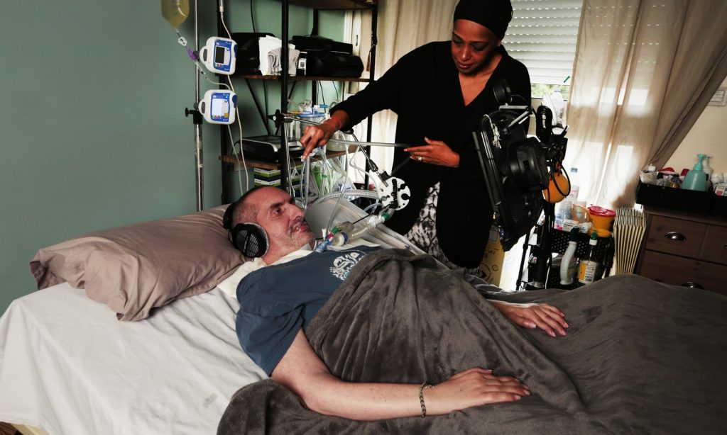 Pone is in bed using eye gaze technology with a computer. His wife is standing next to him feeding a tube in his mouth