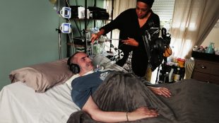 Pone is in bed using eye gaze technology with a computer. His wife is standing next to him feeding a tube in his mouth