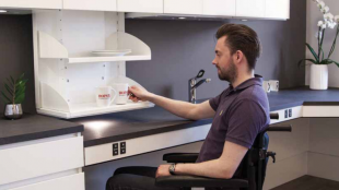 a man in a wheelchair seen grabbing a mug from a kitchen cabinet