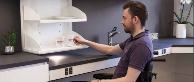 a man in a wheelchair seen grabbing a mug from a kitchen cabinet