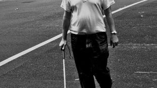 A blind man walking on the road with a white cane in his right hand