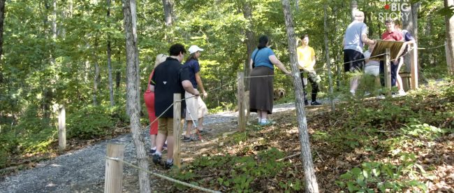 A group of people seen hiking on a Braille trail. They are holding a rope that guides them on the trail.