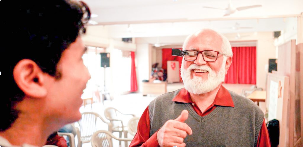 An older man seen interacting with another person and smiling. He is wearing glasses with TranscribeGlass attached to them.