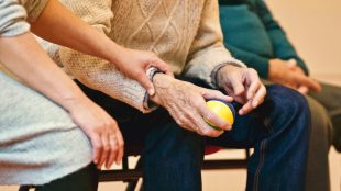 Close up of someone holding the right hand of an elderly person. The elderly person has a yellow ball in their hand.