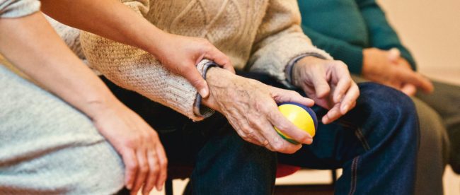 Close up of someone holding the right hand of an elderly person. The elderly person has a yellow ball in their hand.