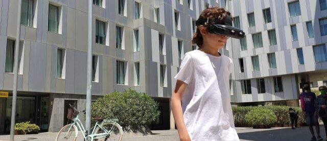 A young boy wearing a white t shirt seen wearing smart glasses outside a building