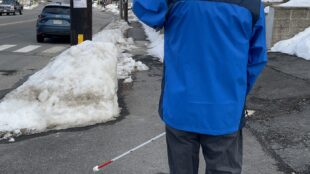 a blind person wearing a blue jacket is seen pointing their phone towards a bus stop sign from a distance