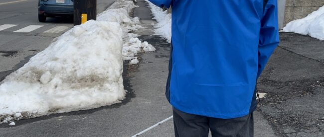 a blind person wearing a blue jacket is seen pointing their phone towards a bus stop sign from a distance