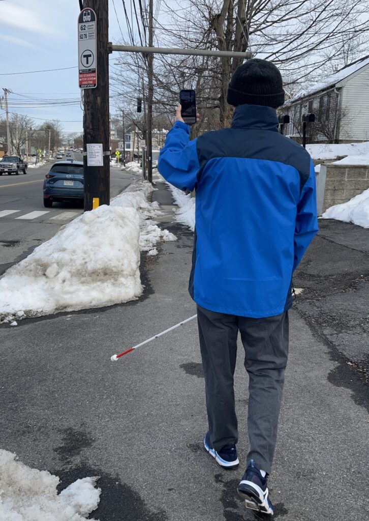 a blind person wearing a blue jacket is seen pointing their phone towards a bus stop sign from a distance