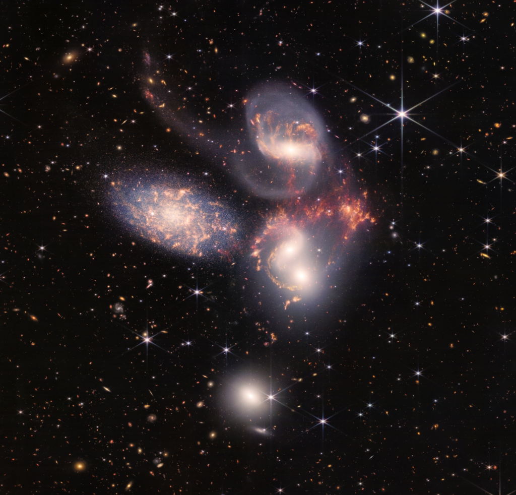 Extended Description
Image of a group of five galaxies that appear close to each other in the sky: two in the middle, one toward the top, one to the upper left, and one toward the bottom. Four of the five appear to be touching. One is somewhat separated. In the image, the galaxies are large relative to the hundreds of much smaller (more distant) galaxies in the background. All five galaxies have bright white cores. Each has a slightly different size, shape, structure, and coloring.
At the center of the image are two bright galaxy cores with orange wisps surrounded by a cloud-like aura of white. The cores are close to each other and there is no clear boundary between the galaxies. The top galaxy of the pair has two spiral arms and the other is more elliptical in shape.
The galaxy toward the top of the image has a bright central core, surrounded by orange wisps and tendrils. The core is surrounded by a thin cloud-like aura of white that forms a diffuse spiral arm-like structures that trails off toward the upper left.
Between the top and center galaxies is a large region of bright orange.
The galaxy toward the bottom of the image sits alone, not appearing to touch any of the other four. It is nearly circular in shape and has a bright core surrounded by a cloud-like aura of white. This galaxy is almost completely white: No orange wisps or arms are apparent.
The galaxy toward the upper left has a fuzzy oval shape, with more distinct points of light than are apparent in the other four galaxies. Within the oval is a bright core with orange wisps and a vague spiral structure. The core of this galaxy is not as bright or distinct as the cores of the other four galaxies.
In the background of the image are numerous smaller, more distant galaxies of various colors, shapes, sizes, and brightness. Scattered across the image, in front of the galaxies are number of foreground stars with diffraction spikes: bright white points, each with eight bright lines radiating out from the center. The sizes of the stars and diffraction spikes vary. Some are superimposed on the large galaxies. The largest is to the upper right of the group of galaxies.
Alt-Text
Image of a group of five galaxies that appear close to each other in the sky: two in the middle, one toward the top, one to the upper left, and one toward the bottom. Four of the five appear to be touching. One is somewhat separated. In the image, the galaxies are large relative to the hundreds of much smaller (more distant) galaxies in the background. All five galaxies have bright white cores. Each has a slightly different size, shape, structure, and coloring. Scattered across the image, in front of the galaxies are number of foreground stars with diffraction spikes: bright white points, each with eight bright lines radiating out from the center.