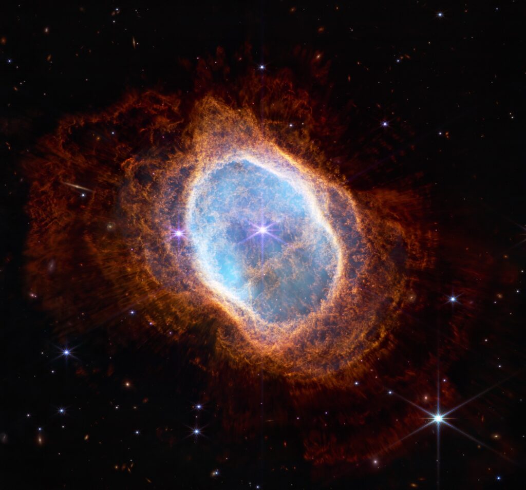 Colorful image of near-infrared light from a glowing cloud with a distorted ring-like shape, illuminated from within by a bright central star. The Southern Ring Nebula is a large, semi-transparent oval that is slightly angled from top left to bottom right. A bright white star appears at the center of this image. A large transparent teal oval surrounds the central star. Several red shells surround the teal oval, extending almost to the edges of the image. The shells become a deeper red with distance from the center. The bright central star has eight diffraction spikes. Behind the gaseous teal layers are deeper orange layers that are arranged like threads in a complex weaving. The red layers, which are wavy overall, look like they have very thin straight lines piercing through them, which are holes where light from a central star is traveling. The background of the image is black and speckled with tiny bright stars and distant galaxies.