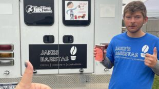 Barista Jake holding a coffee in his right hand and giving a thumbs up with his left. He is wearing a blue t-shirt that says Jake's baristas in larger text and coffee, community, compassion underneath in smaller text. He is posing in front of a truck that has a banner that says "Jakes baristas - coffee, community, compassion). There is also a decal that says "Vinyl closet records" which is father's record store and where the coffee shop is located. THere's another deal that shows a cartoon Jake holding a cup of coffee and the text with barista Jake. There are two dogs at his feet in this cartoon.