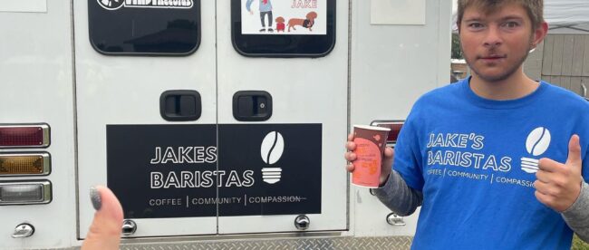 Barista Jake holding a coffee in his right hand and giving a thumbs up with his left. He is wearing a blue t-shirt that says Jake's baristas in larger text and coffee, community, compassion underneath in smaller text. He is posing in front of a truck that has a banner that says 