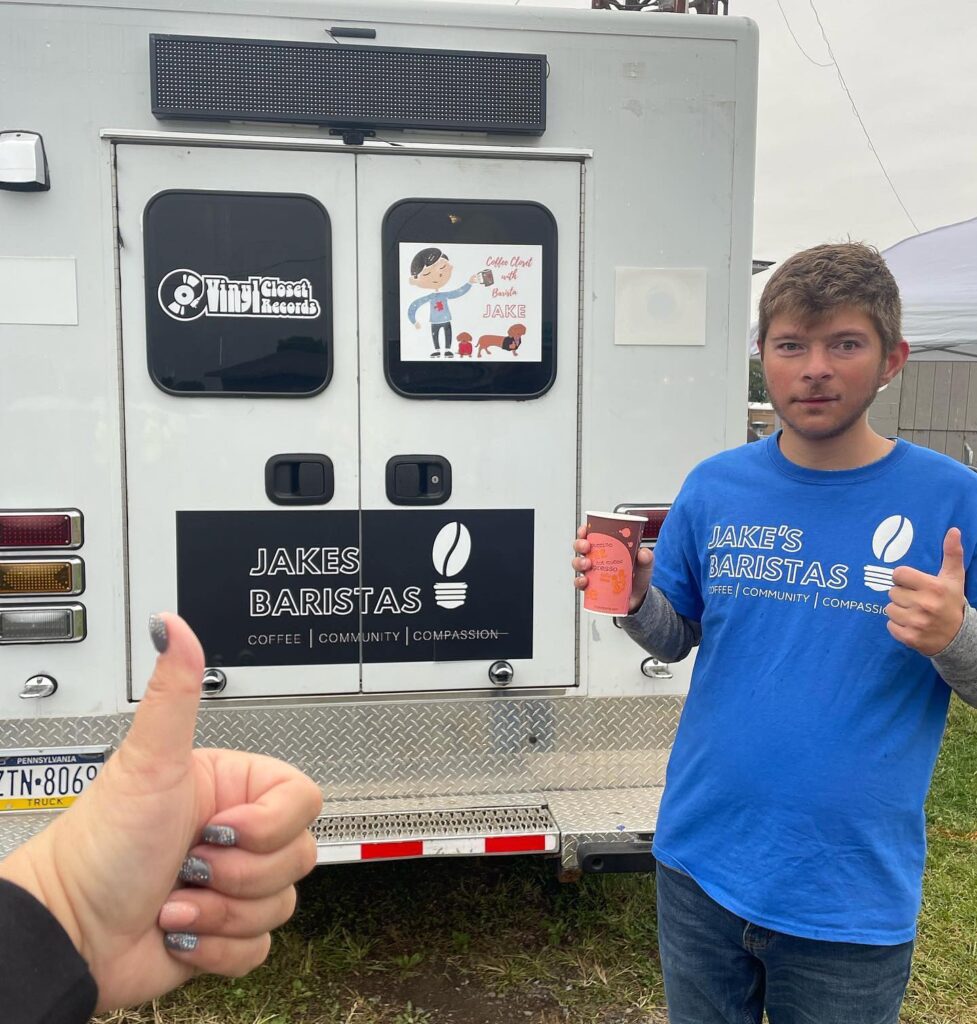 Barista Jake holding a coffee in his right hand and giving a thumbs up with his left. He is wearing a blue t-shirt that says Jake's baristas in larger text and coffee, community, compassion underneath in smaller text. He is posing in front of a truck that has a banner that says "Jakes baristas - coffee, community, compassion). There is also a decal that says "Vinyl closet records" which is father's record store and where the coffee shop is located. THere's another deal that shows a cartoon Jake holding a cup of coffee and the text with barista Jake. There are two dogs at his feet in this cartoon.